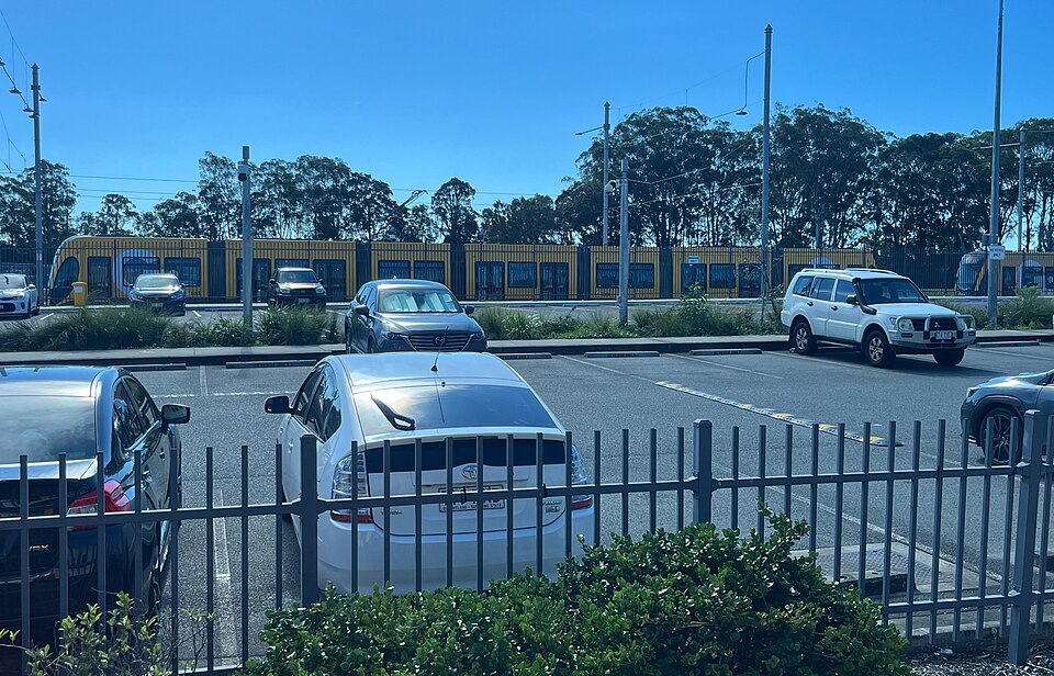Yellow light rail vehicles standing behind a fence at a depot, cars parked in the foreground